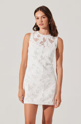 A woman is wearing a sleeveless, white dress with a round neckline. The dress features an intricate lace pattern with geometric shapes and cutouts, and has a delicate scalloped trim along the edges of the armholes, neckline, and hem.